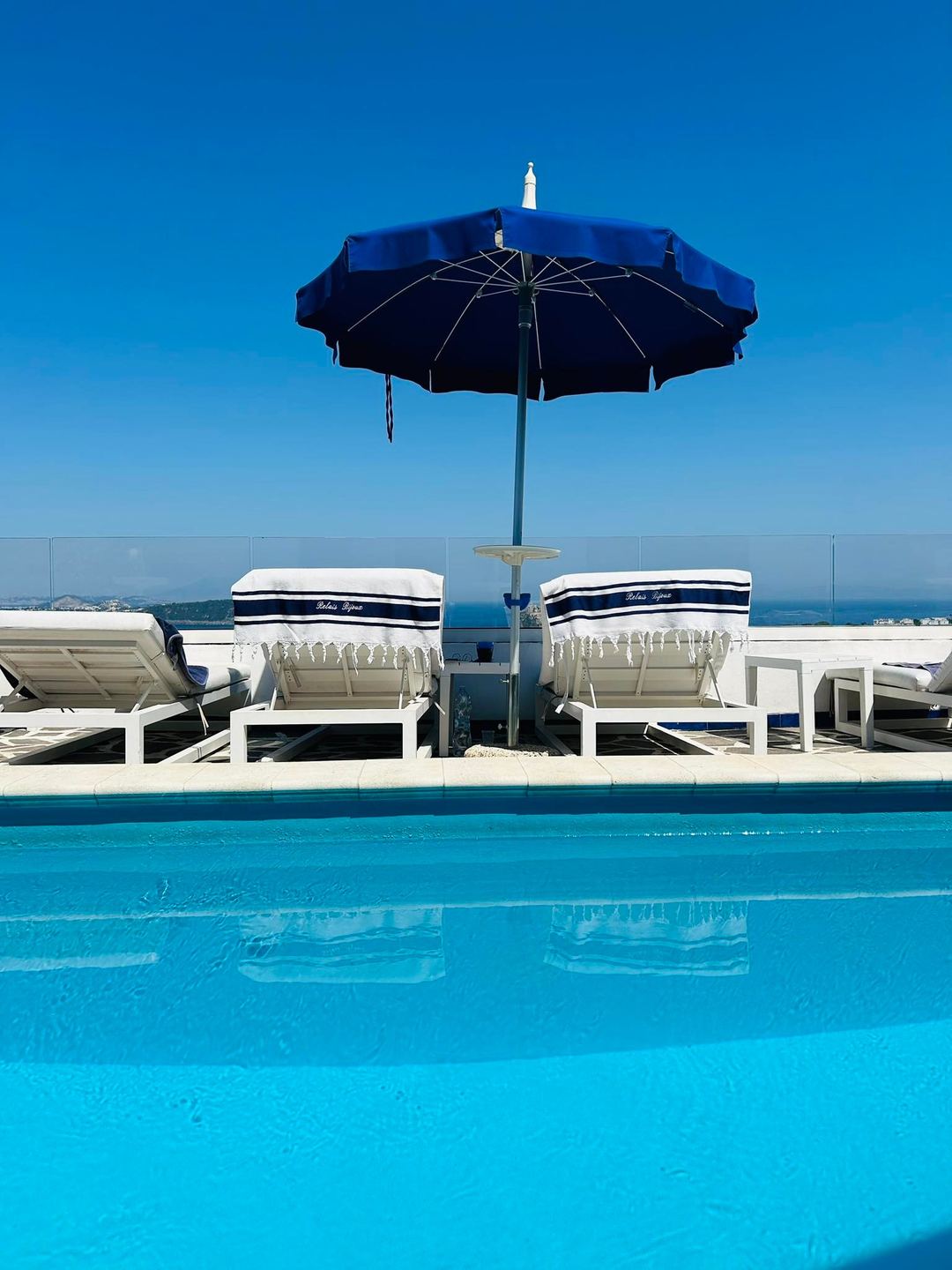 A pristine swimming pool with loungers and a navy blue umbrella overlooking the Mediterranean Sea and coastline under clear blue skies.