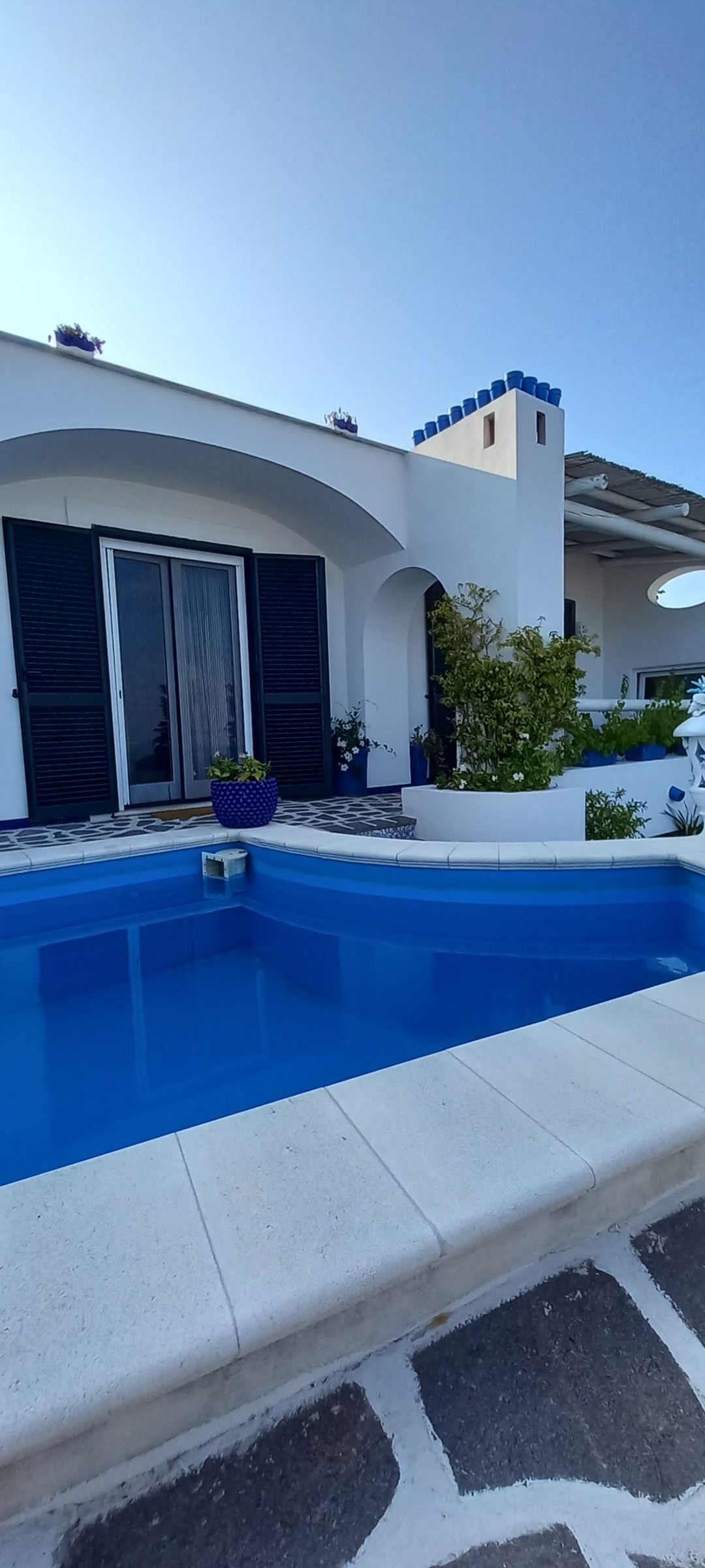 A bright blue swimming pool in front of a modern white villa with curved architecture and dark blue shutters.