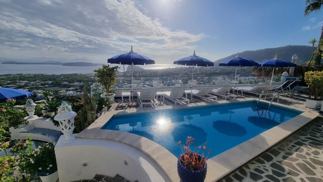 The hotel's swimming pool with blue umbrellas and lounge chairs overlooking the bay and coastal town of Ischia.