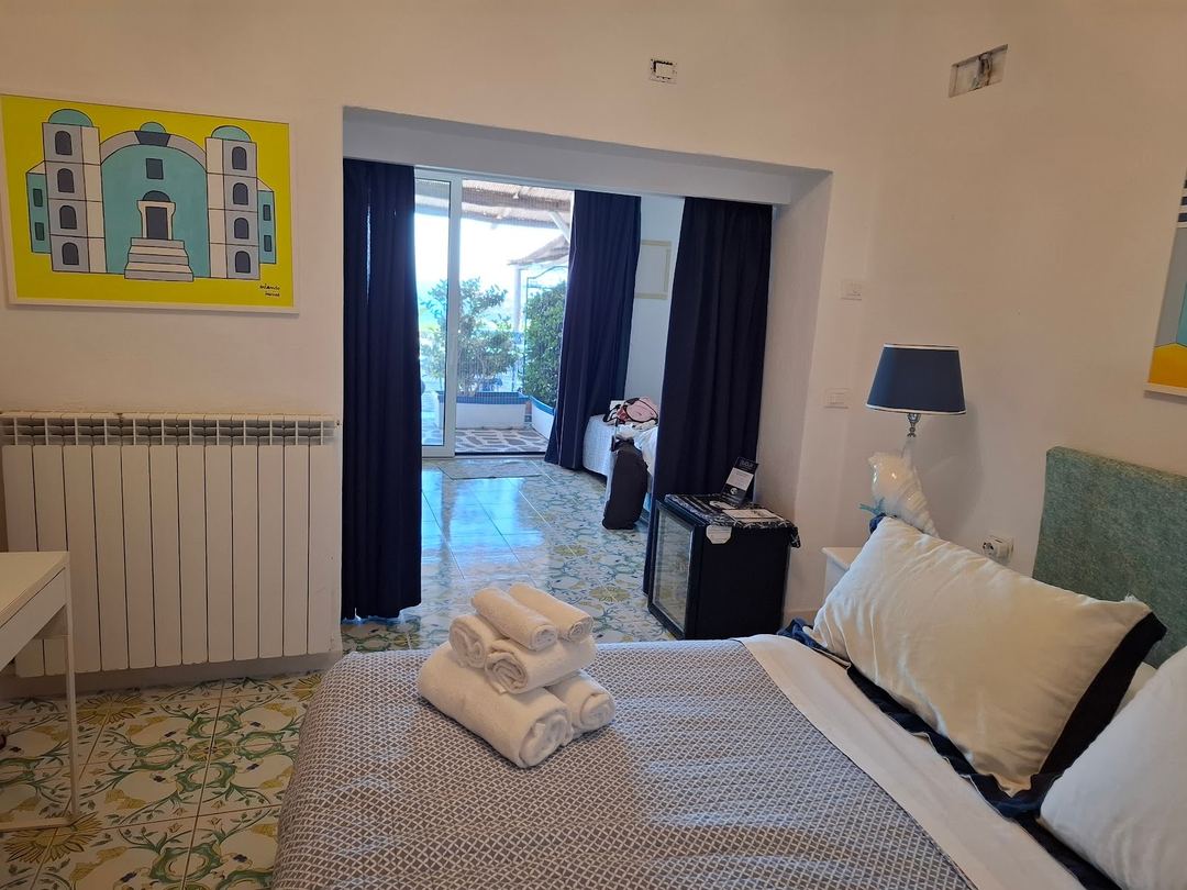 A bright bedroom with contemporary furnishings, featuring a bed with rolled towels, access to a terrace with ocean views, and modern amenities including a desk with lamp and wall radiator.