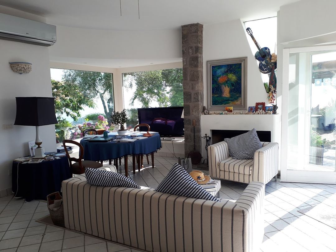 Bright common area with striped upholstered seating, dining table, and views of surrounding greenery through large windows.