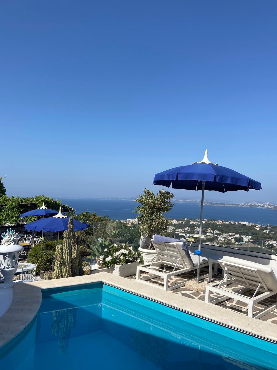 Swimming pool with blue umbrellas and white lounge chairs overlooking the Bay of Naples and Ischia coastline.
