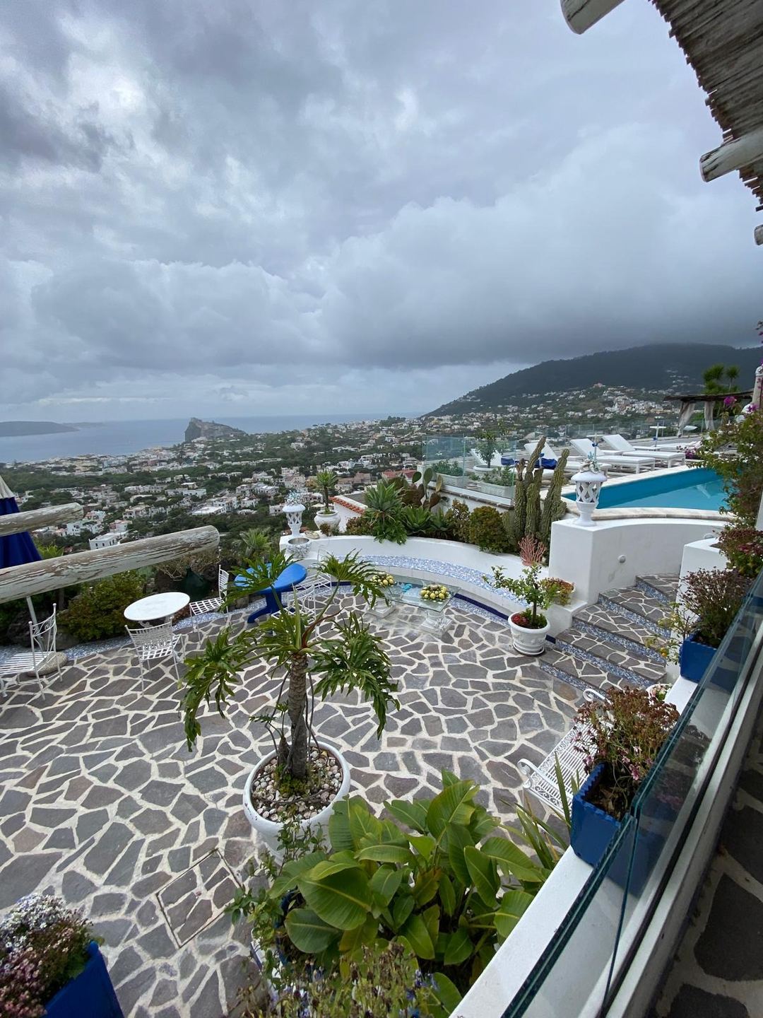 Panoramic view of Ischia's coastline and scattered buildings from an elevated terrace with pool and Mediterranean vegetation.